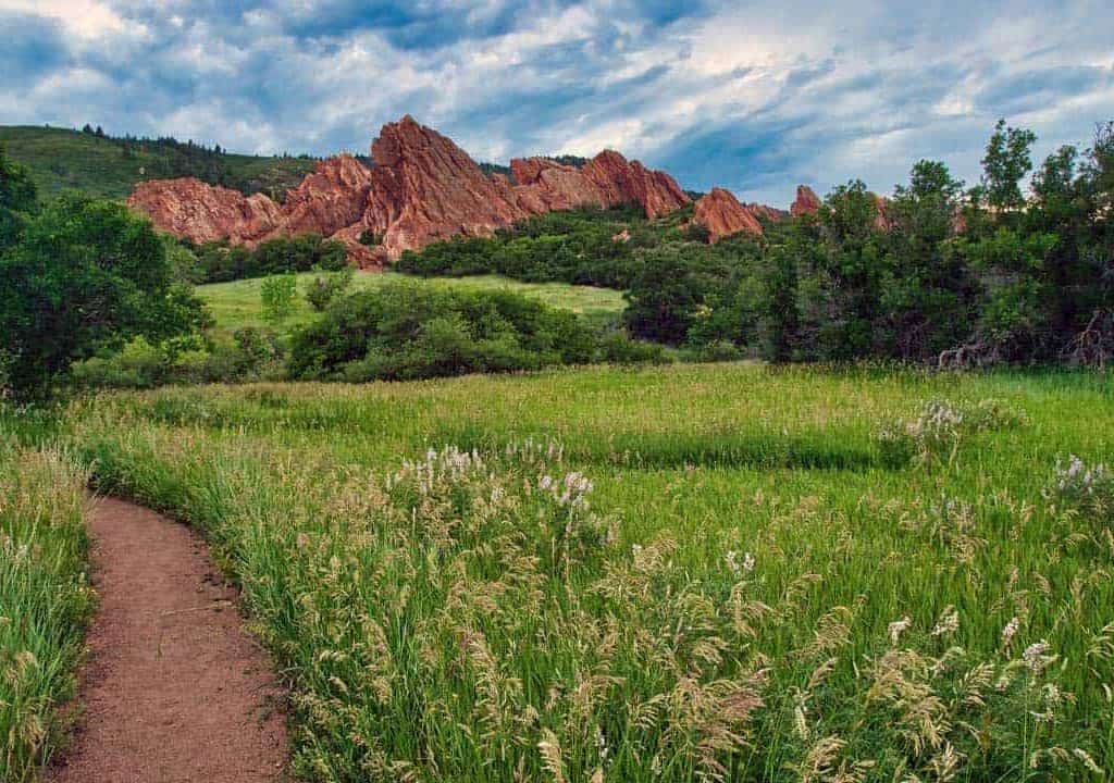 Beautiful green grass and wildflowers line a hiking trail as it winds through the ancient red sandstone fountain formations in Roxborough State Park, Colorado.