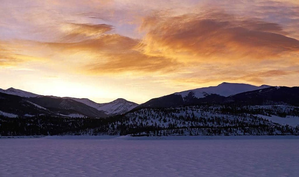 Colorful winter sunrise illuminates the peaks surrounding an icy and frozen Lake Dillon in Summit County, Colorado.