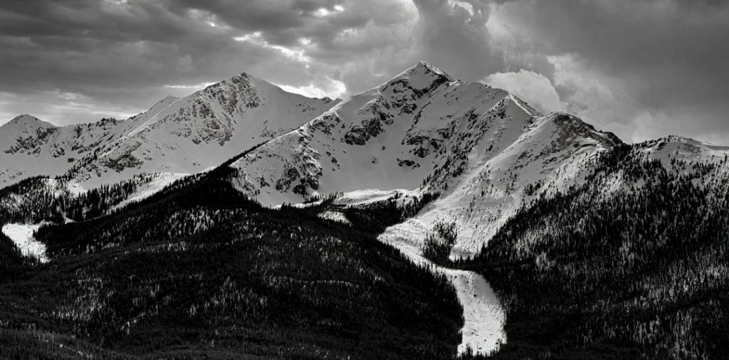 Black and white photograph of clouds rolling in over snow covered Peak One and Ten Mile Peak on a winter afternoon in Summit County Colorado.
