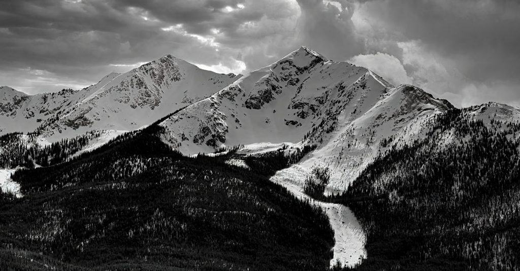 Black and white photograph of clouds rolling in over snow covered Peak One and Ten Mile Peak on a winter afternoon in Summit County Colorado.