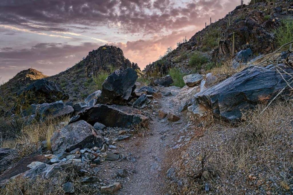 A colorful and moody dawn sky casts soft light over a steep and rocky trail in the Phoenix Mountain Preserve in Arizona.