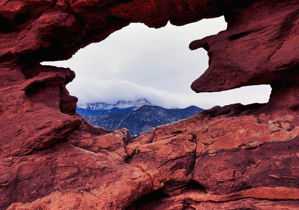 A cloud-capped Pikes Peak seen through a natural rock window in Garden of the Gods park in Colorado Springs, Colorado. The Siamese Twins are two towering rock formations and from the right angle you can catch a uniquely framed view of Pikes Peak through the natural window-like opening between the two rock formations.