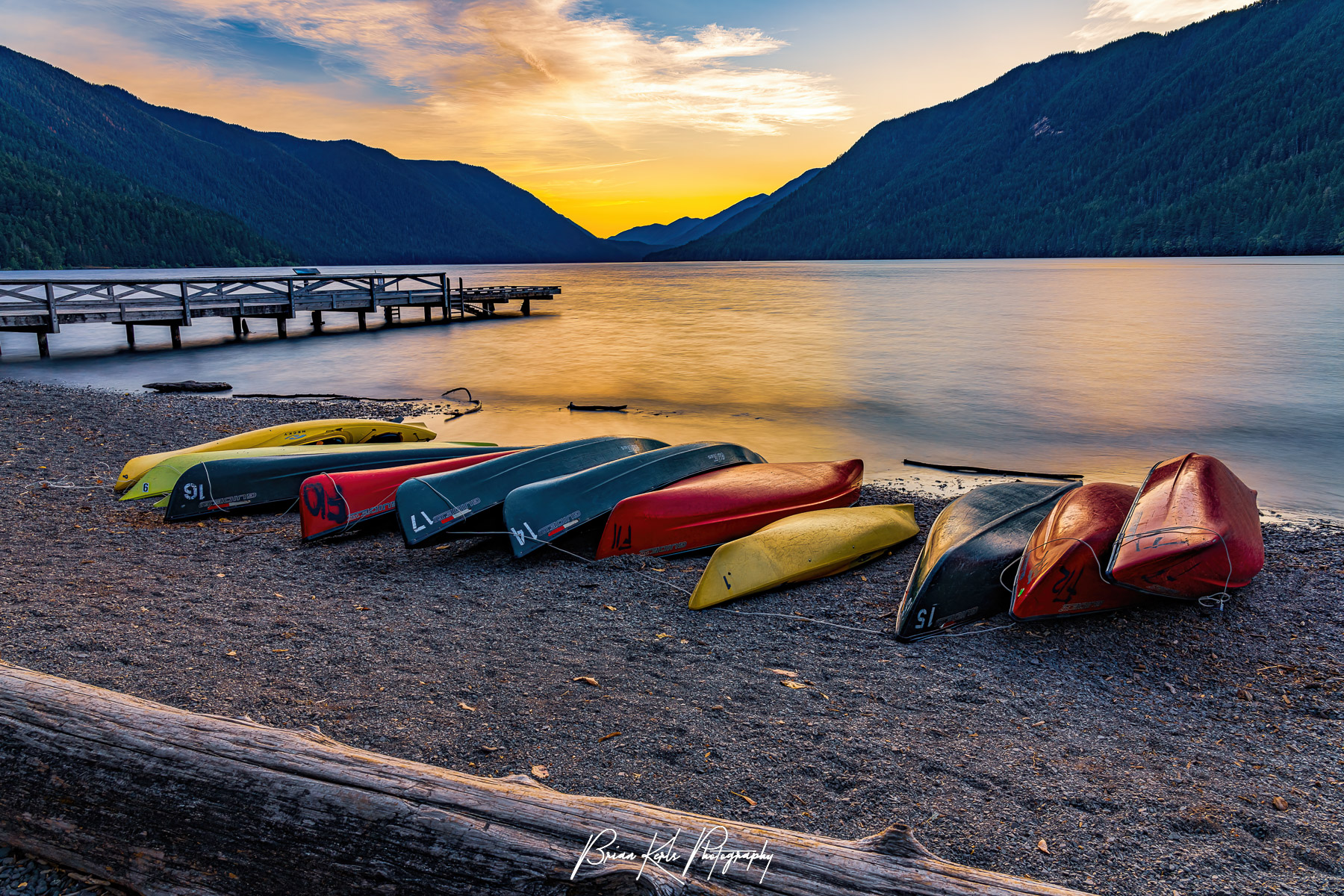 Colorful canoes and kayaks line the shore of Lake Crescent in Olympic National Park in the late evening as sun sets over the calm water and the distant mountains. Nestled in the Olympic Mountains of western Washington the pristine waters of this glacially carved alpine lake glow with the last light of the day on a quiet and peaceful early summer evening.