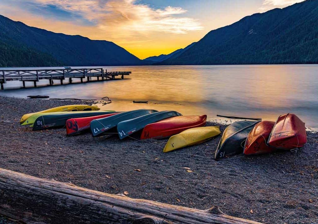 Colorful canoes and kayaks line the shore of Lake Crescent in Olympic National Park in the late evening as sun sets over the calm water and the distant mountains. Nestled in the Olympic Mountains of western Washington the pristine waters of this glacially carved alpine lake glow with the last light of the day on a quiet and peaceful early summer evening.