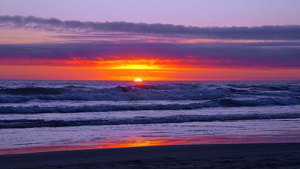 The sun dips below the Pacific horizon casting a colorful sunset glow in the sky and on the crashing surf. Captured on a summer evening while watching the waves of the Pacific Ocean roll onto Cannon Beach on Oregon's North Coast.