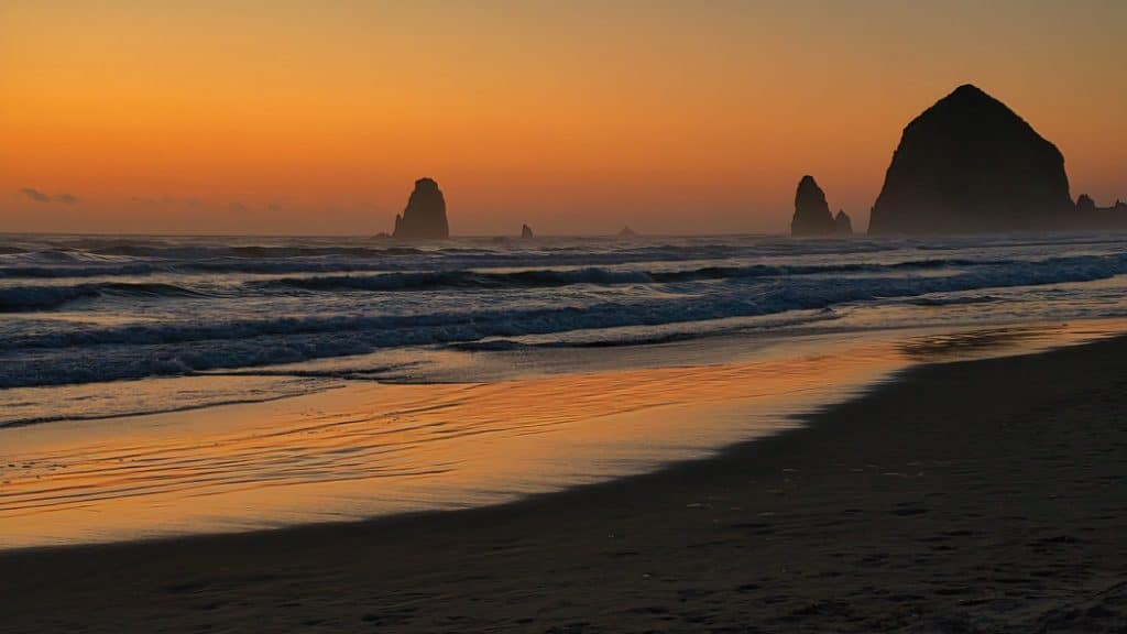 As the sun recedes below the horizon it paints the sky and surf along Cannon Beach, Oregon in vibrant hues of yellow and orange, silhouetting the well-known landmarks of Haystack Rock and The Needles against the colorful sky.