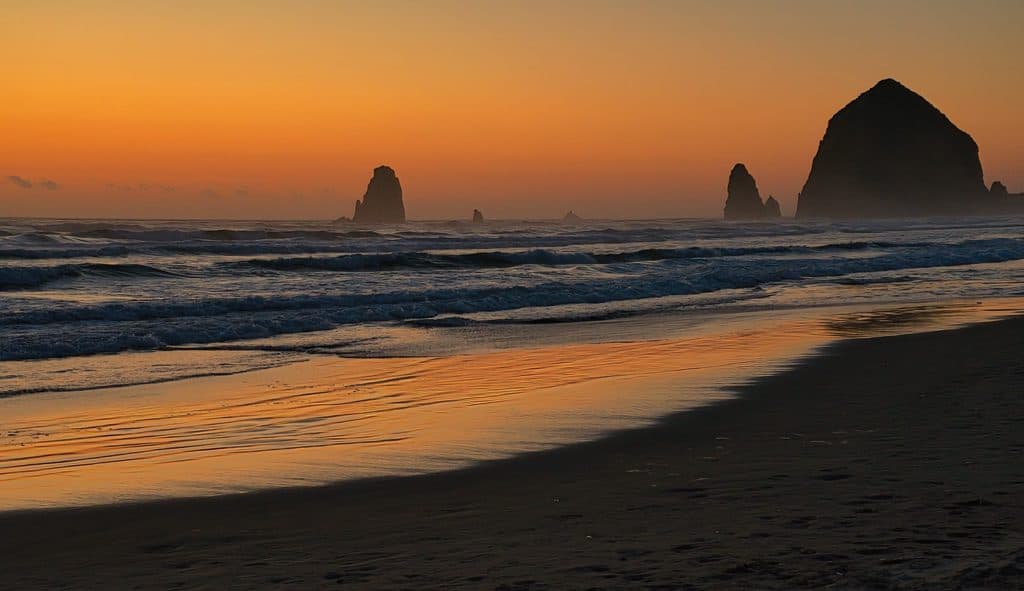 As the sun recedes below the horizon it paints the sky and surf along Cannon Beach, Oregon in vibrant hues of yellow and orange, silhouetting the well-known landmarks of Haystack Rock and The Needles against the colorful sky.