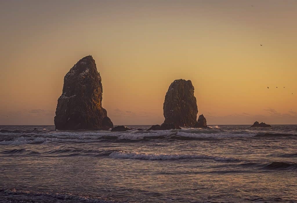 The seastacks known as The Needles standing tall against the crashing waves along Cannon Beach, Oregon as the setting sun paints the sky in delicate hue of pink, yellow, and orange..