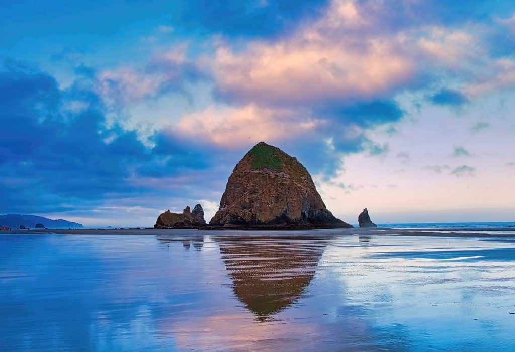 A calm and quiet morning looking south on Cannon Beach, Oregon as the pink, purple, and blue sky of sunrise along the Pacific Coast is reflected in the tidal pools around the iconic Haystack Rock.