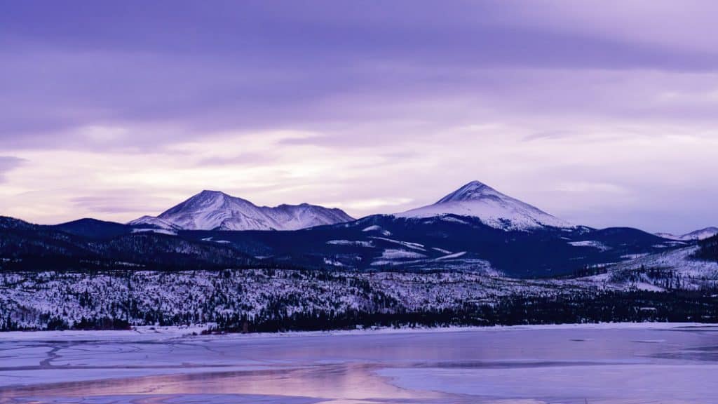 The colorful pink and purple hues of an early winter sunset illuminate the peaks surrounding an icy and partially frozen Lake Dillon in Summit County, Colorado.
