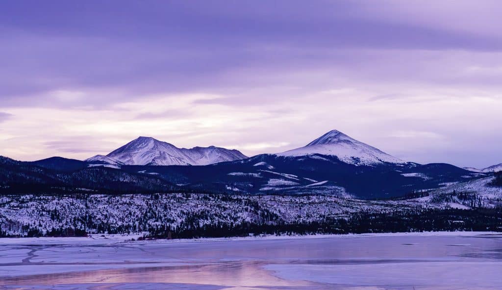 The colorful pink and purple hues of an early winter sunset illuminate the peaks surrounding an icy and partially frozen Lake Dillon in Summit County, Colorado.