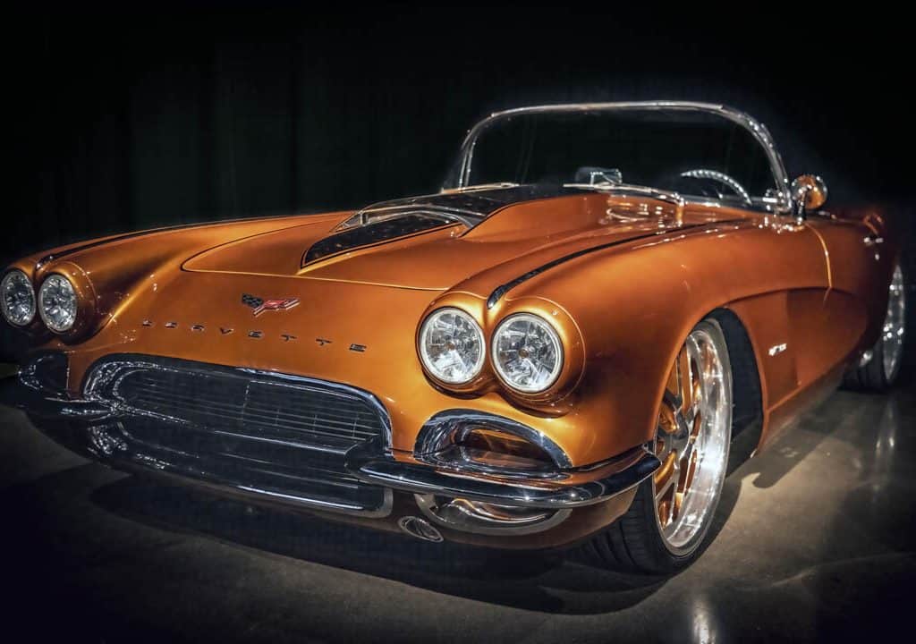 This sleek, customized and restored, vintage 1959 Chevrolet Corvette convertible makes a striking statement with it's gleaming "atomic orange" finish showcased under dramatic lighting. The classic design features round headlights and a chrome bumper, epitomizing timeless automotive elegance.