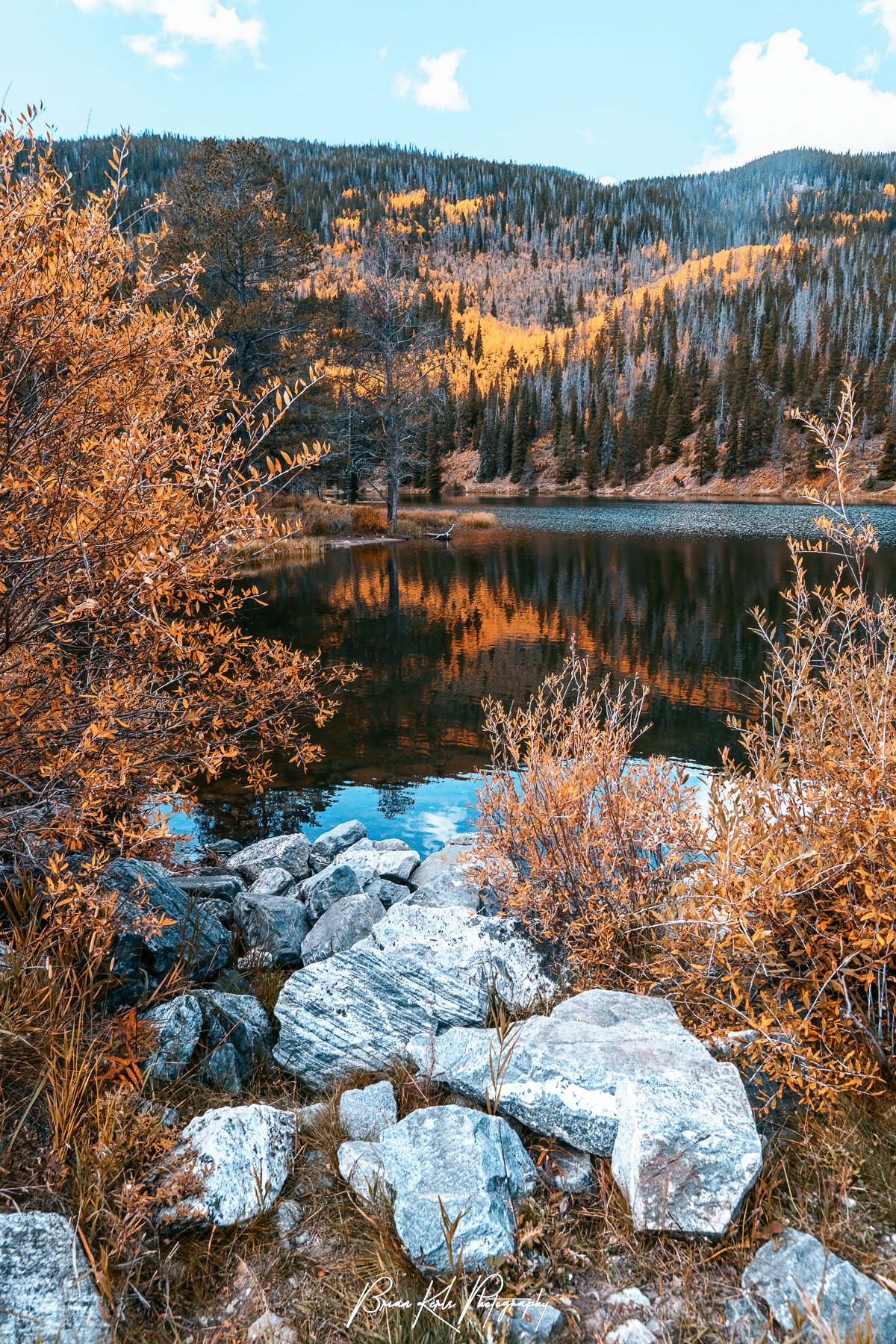 Bands of remaining fall color mixed in with the evergreen forest are reflected in the moody water of Officer's Gulch pond in Summit County, Colorado on an early morning in late autumn.