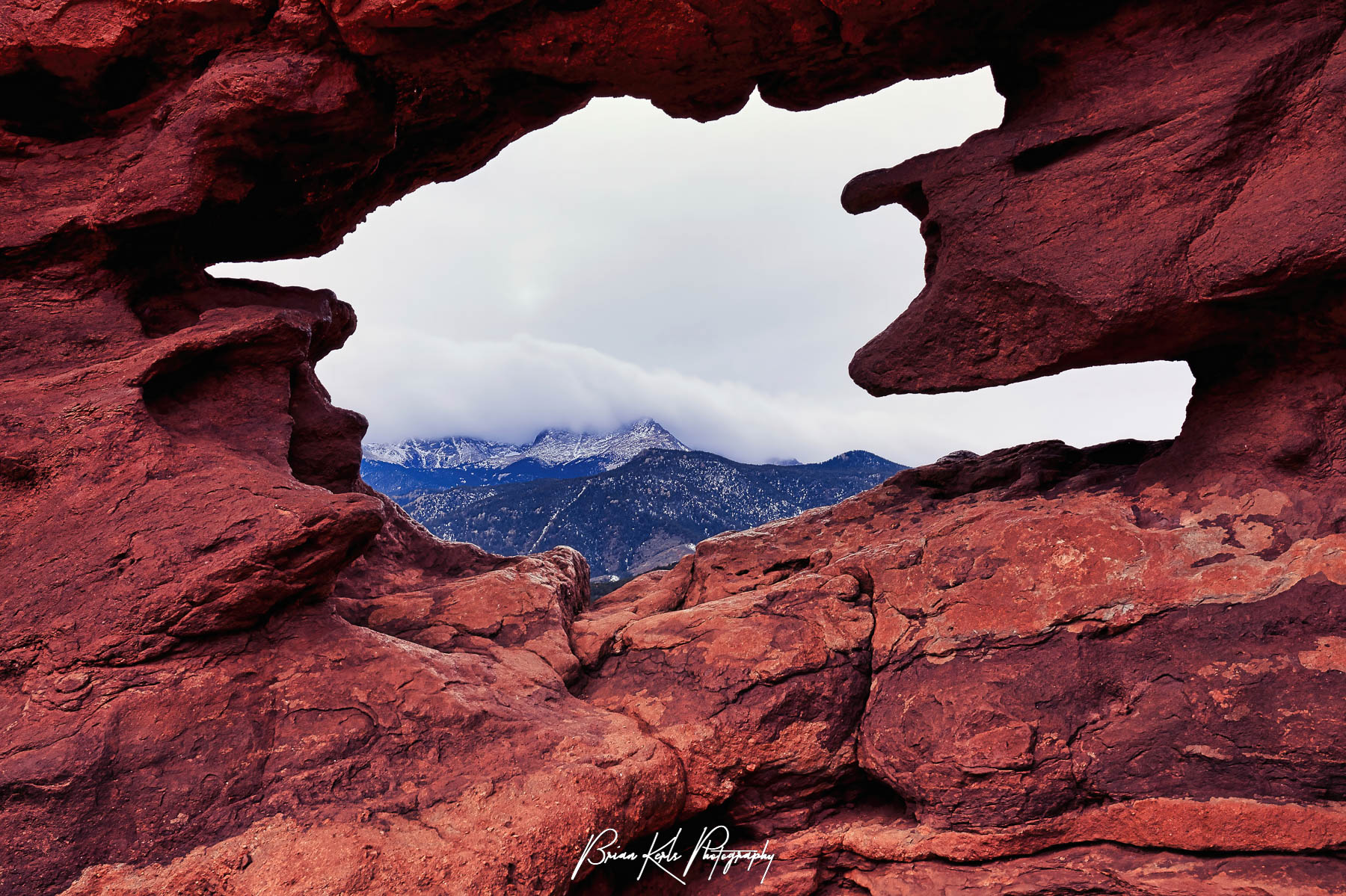 A cloud-capped Pikes Peak seen through a natural rock window in Garden of the Gods park in Colorado Springs, Colorado. The Siamese Twins are two towering rock formations and from the right angle you can catch a uniquely framed view of Pikes Peak through the natural window-like opening between the two rock formations.