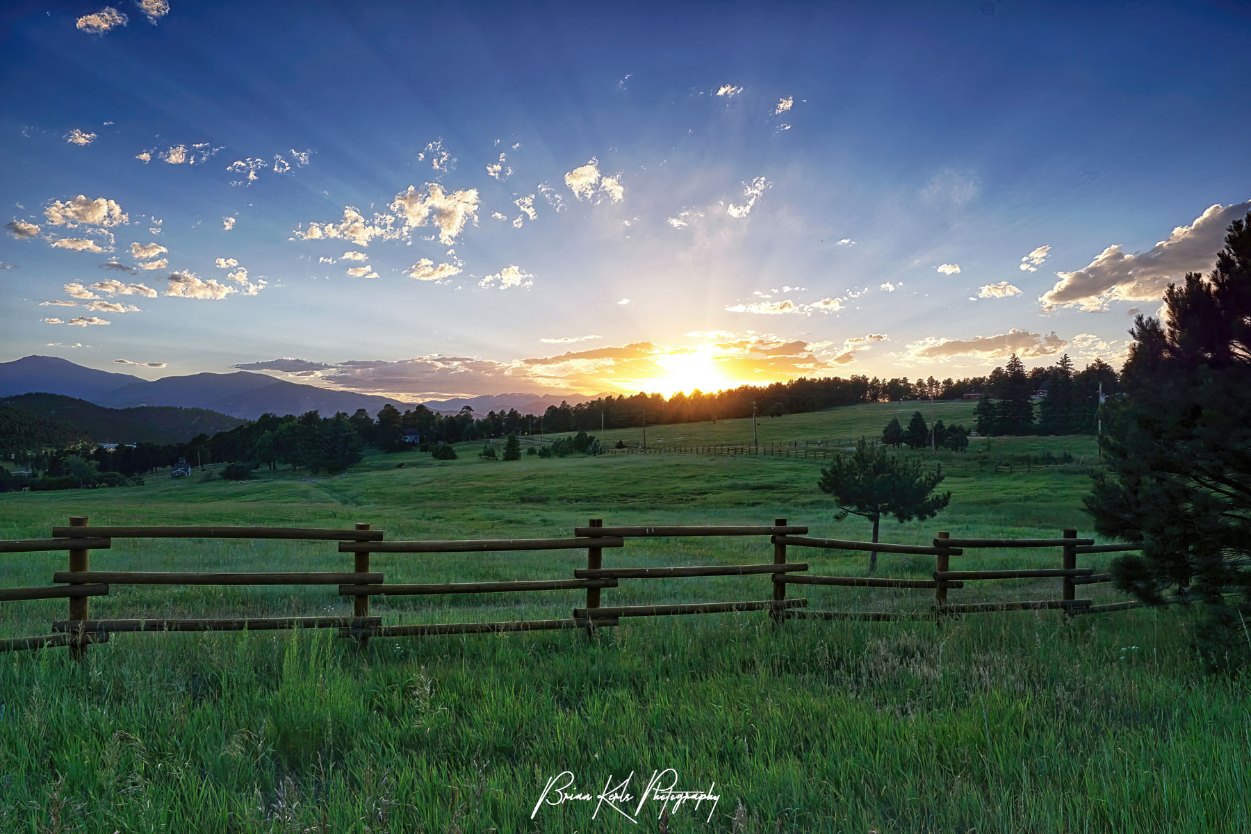 Rays streak the sky over a lush green meadow as the setting sun slips beyond the horizon. This image was captured on a warm summer evening in the foothills of the Rocky Mountains near Genesee, CO.