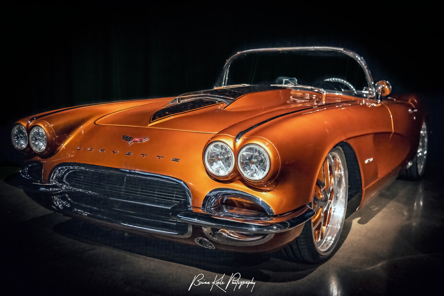 This sleek, customized and restored, vintage 1959 Chevrolet Corvette convertible makes a striking statement with it's gleaming "atomic orange" finish showcased under dramatic lighting. The classic design features round headlights and a chrome bumper, epitomizing timeless automotive elegance.