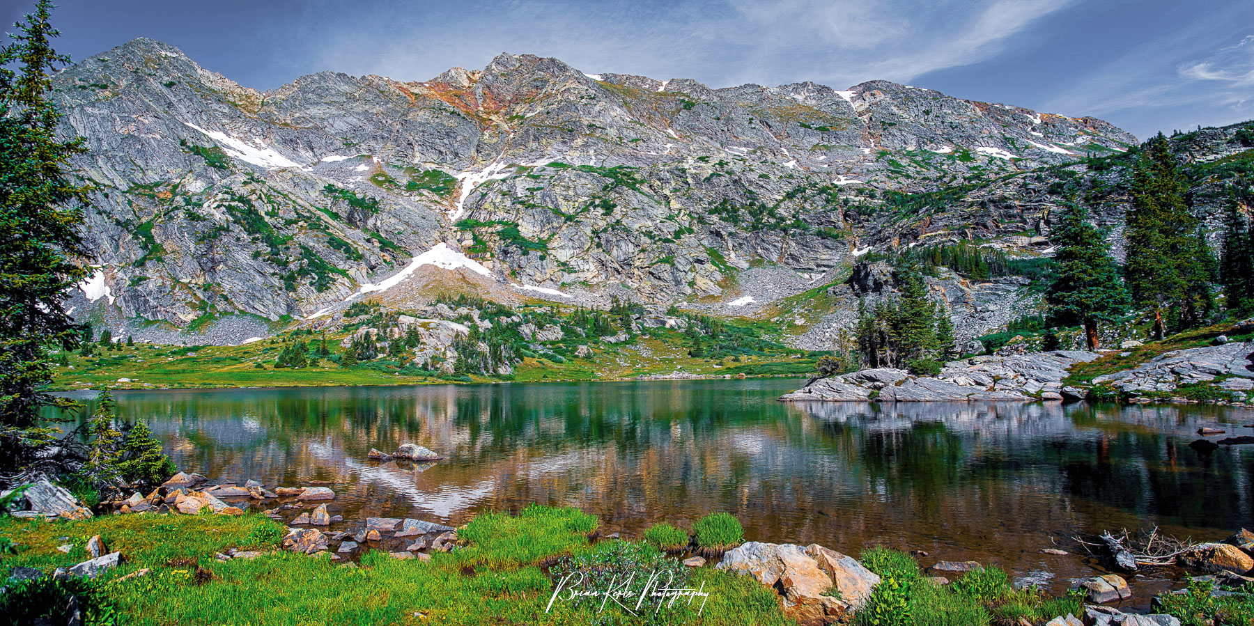 Panoramic view looking across the lush green shoreline of Lower Missouri Lake toward the rugged ridgeline of 12,000' peaks that tower over it in the Holy Cross Wilderness of Colorado.