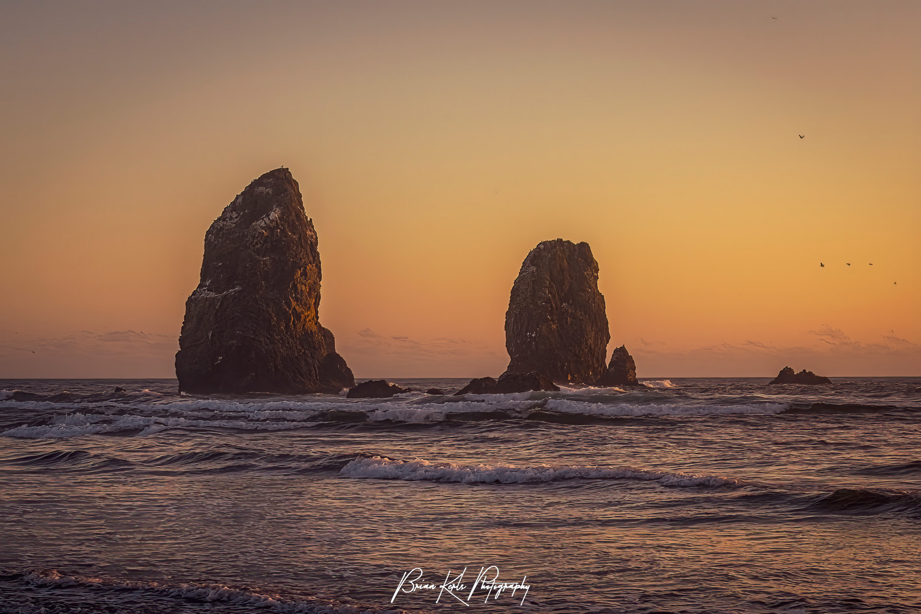 The seastacks known as The Needles standing tall against the crashing waves along Cannon Beach, Oregon as the setting sun paints the sky in delicate hues of pink, yellow, and orange.