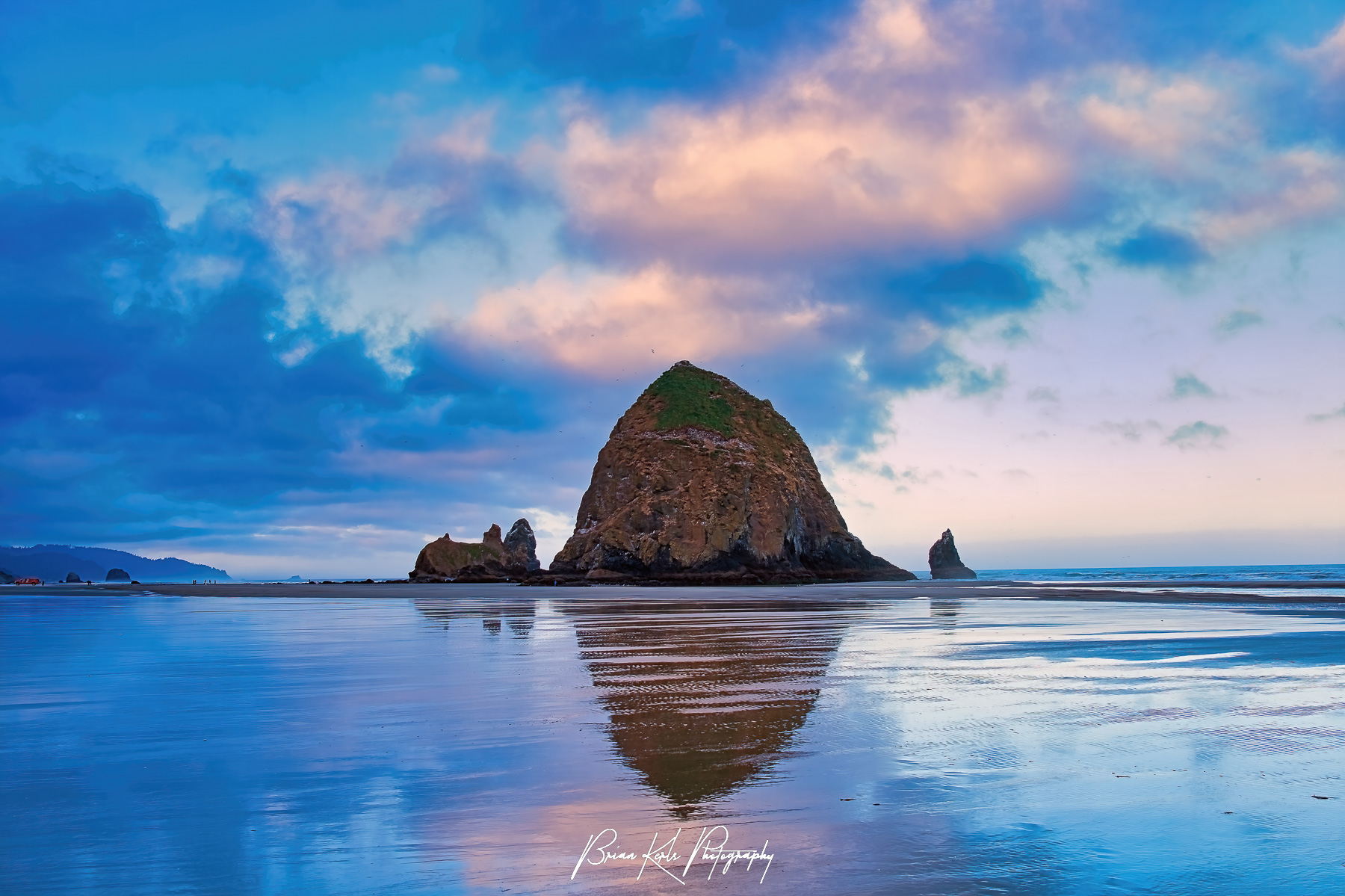 A calm and quiet morning looking south on Cannon Beach, Oregon as the pink, purple, and blue sky of sunrise along the Pacific Coast is reflected in the tidal pools around the iconic Haystack Rock.