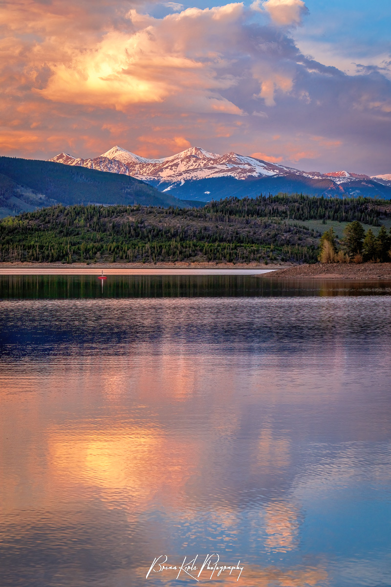 Grays and Torreys Peaks glow beneath the billowing pink and orange clouds of a dramatic spring sunset reflected in the water of Lake Dillon in Summit County, Colorado.