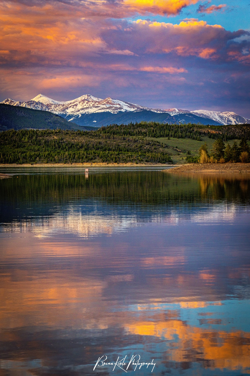 Snow-capped Grays and Torreys Peaks stand silently, glowing in the last light of the day, as the colorful spring sunset drenches the billowing clouds and paints Lake Dillon with their reflections.