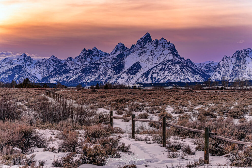 Colorful winter sunset sky over the snow capped peaks of the Grand Tetons with rustic fence and field of sagebrush in foreground.
