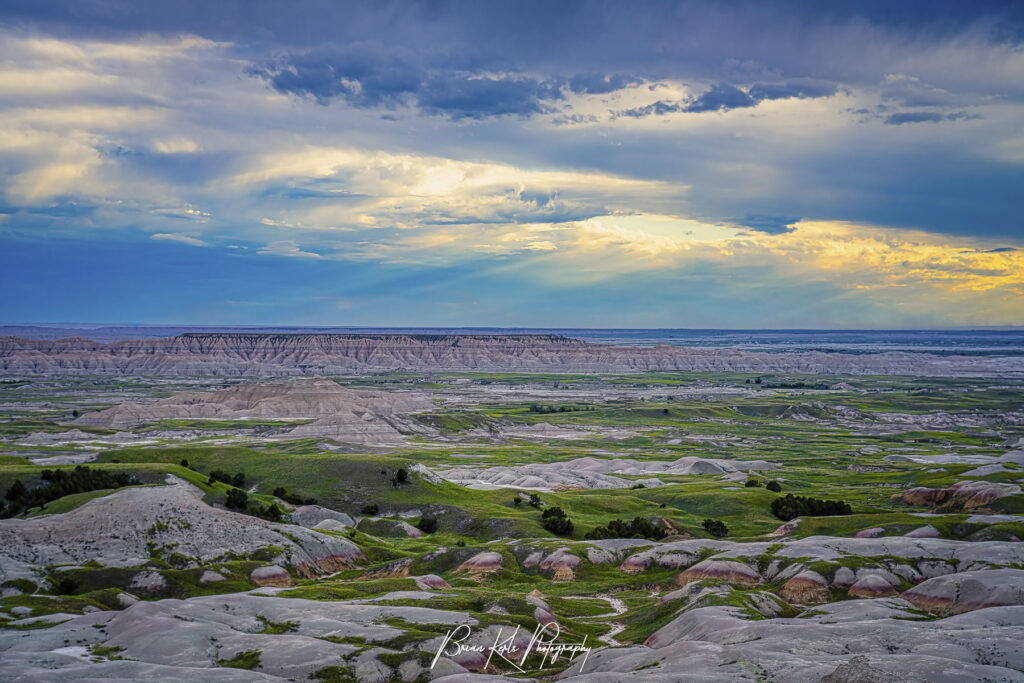 Wide view of layered Badlands National Park ridges with green patches under a dramatic blue sky with clouds, at dusk/early evening.