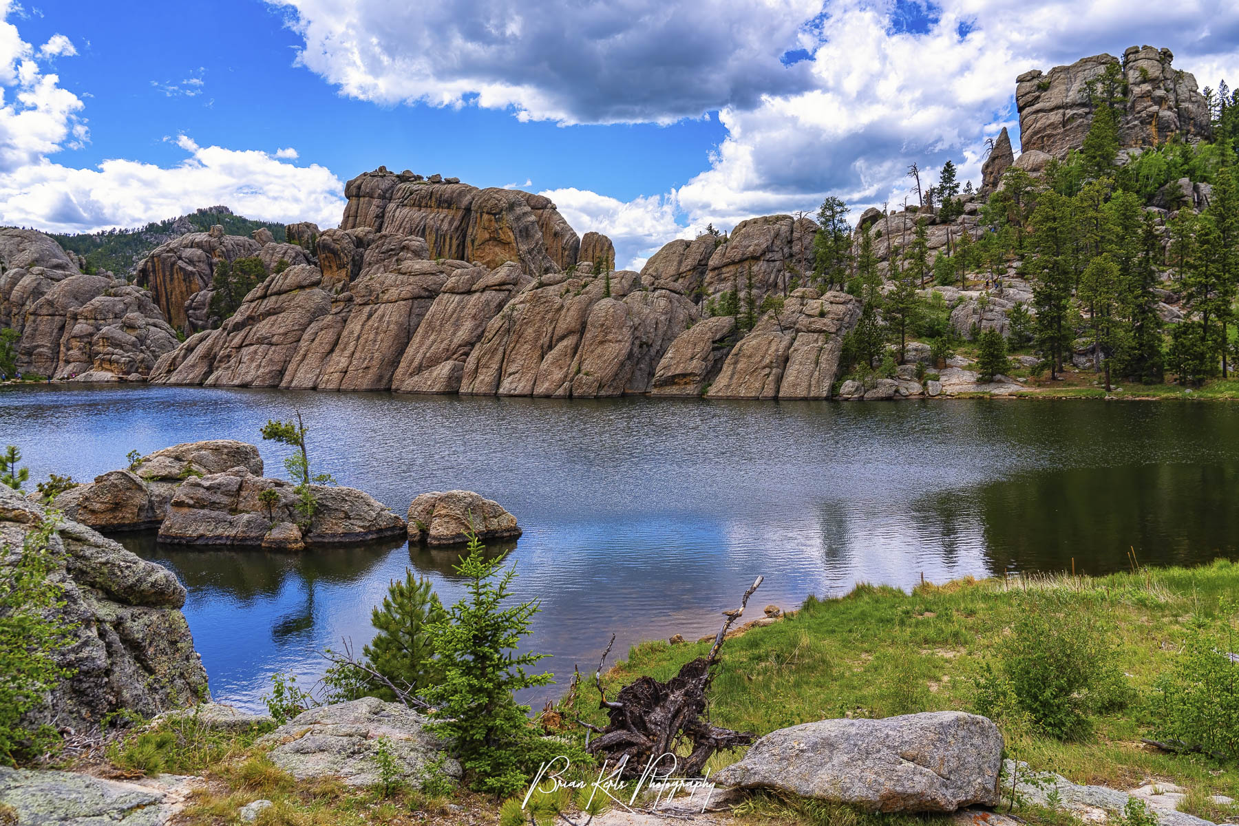 Scenic Sylvan Lake surrounded by large granite rock formations and pine trees under a blue sky with scattered clouds, calm water reflecting the landscape.